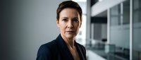 Middle-aged woman with short dark hair and fair complexion wearing navy blazer in modern office environment with glass partitions and neutral background.