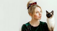 Young woman with blonde hair styled in updo with burgundy bow, wearing green velvet top, posing with Siamese cat against white background for portrait and lifestyle use.