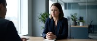 Professional businesswoman in navy blazer during corporate meeting or job interview in modern office with natural lighting and plants.