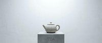 Elegant white ceramic teapot with textured surface displayed on a concrete pedestal with TEA inscription against a soft gray background.