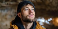 Young bearded Caucasian man in yellow jacket and knit hat looking up thoughtfully in urban tunnel setting with warm bokeh lights in background.