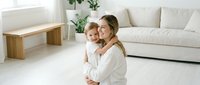 Young blonde mother holding smiling baby in bright modern living room with white sofa and wooden furniture for family lifestyle content.