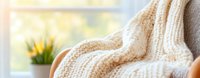 Cozy knitted beige blanket draped over armchair near window with yellow daffodils in pot, creating warm and inviting home atmosphere. Soft natural lighting adds comfort.