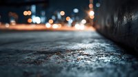 Urban sidewalk at night with blurred city lights in background, creating atmospheric mood with textured concrete surface.