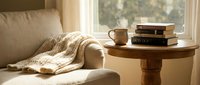 Cozy reading corner with knitted blanket on beige sofa, coffee cup and books on wooden table by sunny window for relaxation and leisure lifestyle.