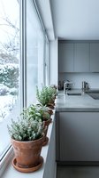Modern minimalist kitchen with herb garden on windowsill overlooking snowy winter landscape. Fresh potted plants bring natural elements to contemporary interior design.
