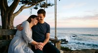 Young couple embracing on wooden swing by ocean at sunset, romantic moment together on coastal cliff with tree, peaceful evening light and waves below.