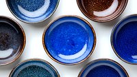 Handcrafted ceramic bowls in shades of deep blue and brown with speckled glazed texture arranged on white background, top view composition for culinary styling.