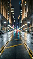 Wet urban street at night with glowing bokeh city lights, tall buildings, and yellow road markings reflecting on rain-soaked asphalt.