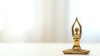 Golden bronze meditation figurine in lotus pose with raised arms on white surface against soft blurred background for wellness and mindfulness concepts.
