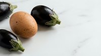 Fresh organic eggplants and brown chicken egg arranged on white marble surface for healthy cooking and farm-to-table cuisine preparation.