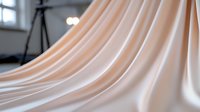 Elegant peach-colored silk fabric with soft flowing folds and gentle waves creates luxurious drapery texture against neutral background, suitable for fashion and interior design.