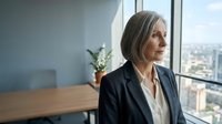 Mature woman with gray hair wearing black blazer and white blouse, looking out floor-to-ceiling window in modern high-rise office with cityscape view, professional business environment.