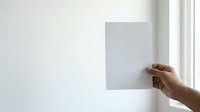 Person with medium skin tone holding blank white paper against white wall, suitable for mockups, presentations, advertising campaigns, and design templates.