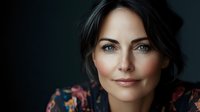 Confident middle-aged woman with blue eyes and dark hair smiling gently at camera against dark background, wearing floral pattern blouse. Professional portrait with soft lighting.