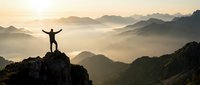 Silhouette of a man standing on a rocky mountain peak at sunrise, arms raised in triumph, with misty valleys and layered mountain ranges below.