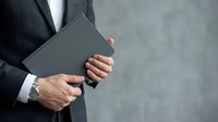 Businessman in dark suit holding tablet device against gray concrete wall background, professional technology concept for corporate business and digital solutions.