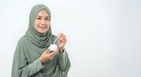Young Muslim woman in green hijab holding skincare cream jar, smiling at camera on white background for beauty and cosmetics advertising.