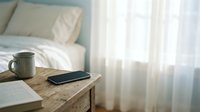 Cozy bedroom interior with smartphone and coffee mug on wooden nightstand beside unmade bed with white linens and soft natural window light.