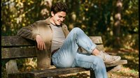 Young Caucasian man in beige jacket relaxing on wooden park bench during golden hour autumn day with warm sunlight filtering through trees.