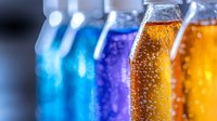 Colorful carbonated drinks in glass bottles with bubbles and fizz, arranged in row showing gradient from blue to orange, refreshing summer beverages in macro view.