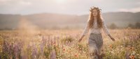 Young woman with long curly reddish-brown hair wearing a lace veil and flowing dress standing in a wildflower meadow during golden hour, surrounded by purple and white blooms with misty mountains in the background.