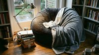 Cozy reading nook with comfortable armchair draped in soft blanket, stacked books, warm lighting, and window view. Perfect for relaxation, home decor, and lifestyle content.