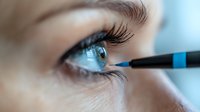 Close-up view of blue eye with long eyelashes and eyebrow, black eyeliner pencil applying makeup. Macro beauty shot with soft focus background.