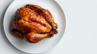 Whole roasted golden brown chicken with crispy skin served on white round plate against light background, top view food photography.