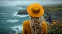 Young woman in yellow sun hat and sweater looking at dramatic coastal cliffs and stormy ocean waves, wind blowing through long blonde wavy hair, back view perspective.