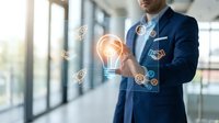 Professional businessman in dark suit holding glowing lightbulb with digital innovation icons in modern office environment representing creative business solutions.
