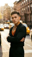 Confident young Hispanic businessman in black suit and patterned scarf standing with crossed arms on urban city street during golden hour sunset.