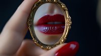 Close-up of bright red lips reflected in vintage gold ornate hand mirror against dark background, with matching red manicured nail visible.