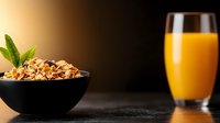 Breakfast cereal in black bowl with fresh mint leaf and glass of orange juice on dark surface with warm lighting.