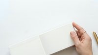 Adult hand holding open blank notebook with cream pages against neutral background for branding, stationery mockup, or editorial layouts.