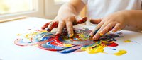 Child hands creating colorful finger painting artwork on white paper with vibrant red, blue, yellow paint smears in bright indoor setting.
