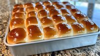 Fresh baked dinner rolls with golden brown tops arranged in rows on metal baking pan, showing soft texture and glossy butter-brushed surface.
