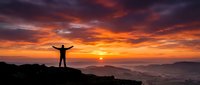 Silhouette of person with raised arms celebrating success on mountain peak at dramatic sunrise with orange and purple clouds over layered hills.