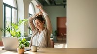 Young Asian businesswoman stretching arms above head during work break at modern office desk with laptop and coffee cup for wellness and productivity.