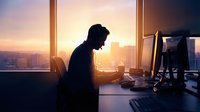 Young professional businessman working late at computer in modern office with city skyline view during golden hour sunset creating productive workspace atmosphere.