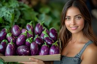 Young Caucasian woman in denim overalls holding wooden crate with fresh purple eggplants, smiling at camera against green garden background. Organic farming concept.