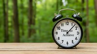 Vintage alarm clock with motivational text "Time to Achieve" on wooden table against blurred green forest background, symbolizing personal growth and success.