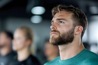 Confident athletic Caucasian male with beard wearing green t-shirt looking up with determination during workout session in modern fitness center. Blurred people in background.