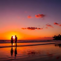 Romantic couple silhouettes holding hands on tropical beach at sunset with golden sky reflections on wet sand for vacation and travel concepts.