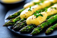 Fresh green asparagus spears with melted hollandaise sauce and black pepper served on dark plate, macro food photography with selective focus.