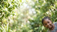 Young African girl smiling outdoors surrounded by lush green foliage and natural sunlight, expressing joy and happiness in nature setting for lifestyle and wellness content.