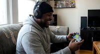 Young African American man wearing headphones playing mobile games on smartphone in modern living room with entertainment setup.