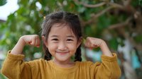Cheerful Asian girl with braided hair in yellow sweater showing strength by flexing arms outdoors against blurred green nature background.