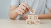 Business person building wooden blocks with strategy icons including target, lightbulb, gear, growth chart and team symbols for planning and development.