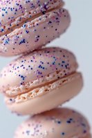 Pink macarons with blue and purple sprinkles arranged in diagonal composition, close-up view showing delicate texture and filling details on white background.